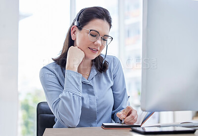 One young caucasian call centre telemarketing agent wearing glasses and writing notes while talking on a headset and working on a computer in an office. Female consultant operating helpdesk for customer service and sales support