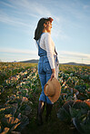 Woman farmer standing in a cabbage field on a farm. Young female with a straw hat and rubber boots looking over her field of organic vegetables