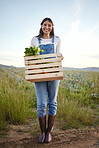 Portrait of a woman farmer holding a wooden box of fresh vegetables. Young brunette female standing on a field while holding a basket of organic produce