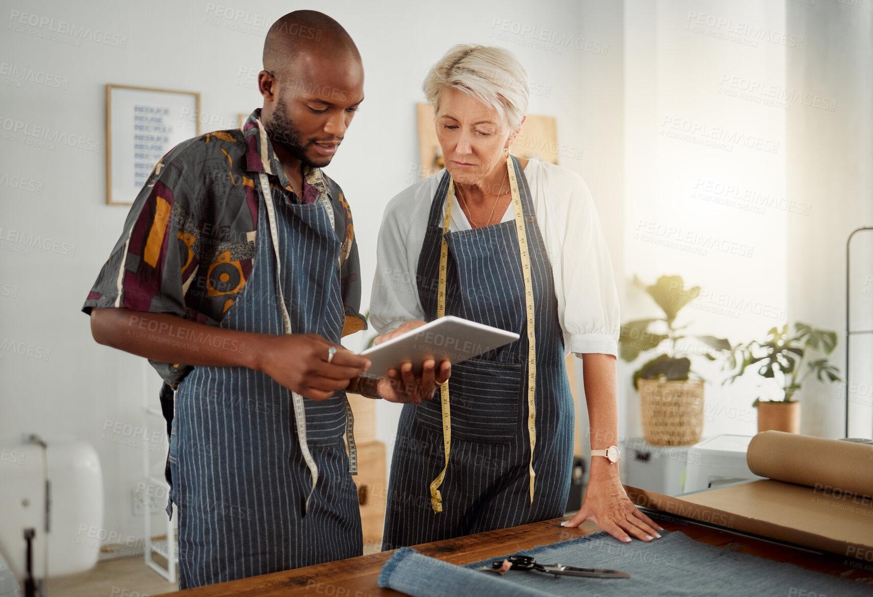 Buy stock photo Two designers using a digital tablet while working with material at work. Young african american tailor talking and holding a digital tablet while working with a female caucasian colleague