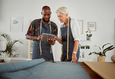 Buy stock photo Two clothing designers using a digital tablet while working with material at work. Young african american tailor talking and holding a digital tablet while working with a female caucasian colleague