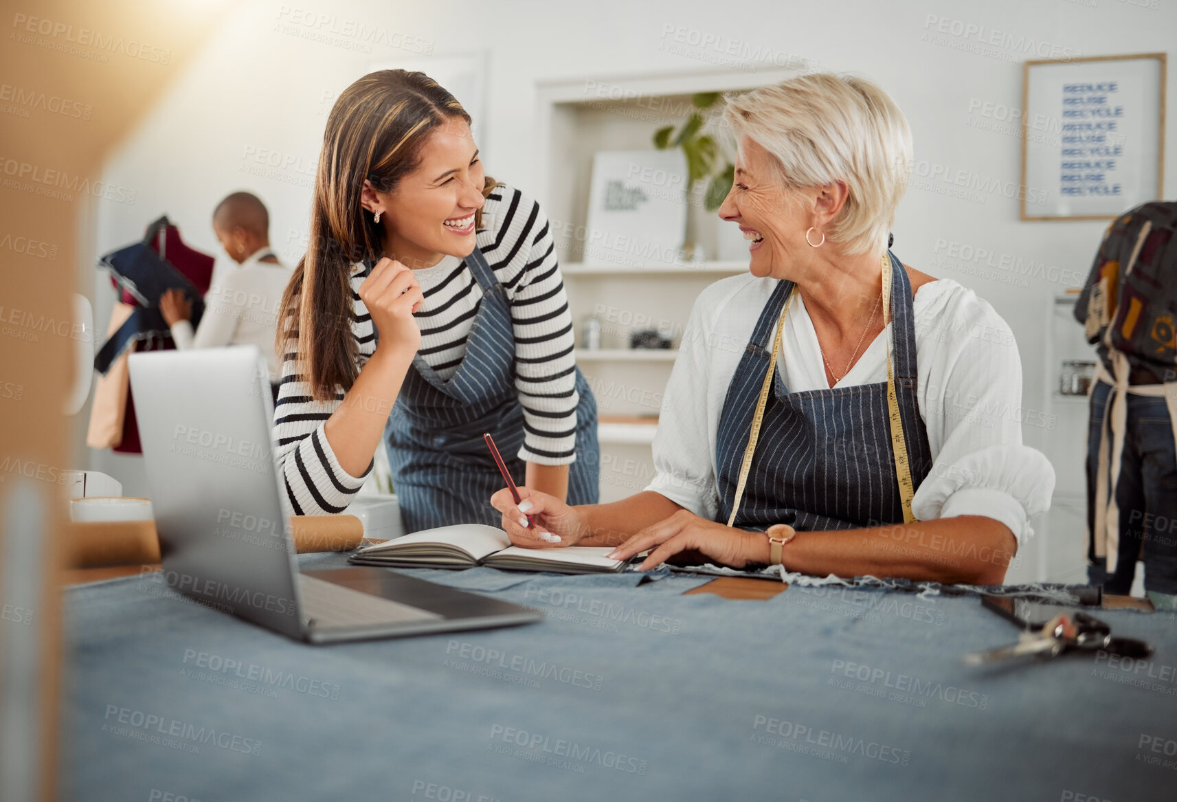 Buy stock photo Two cheerful clothing designers using a laptop and writing in a notebook while working with material in a meeting at work. Caucasian and mixed race female tailors talking and laughing together at work