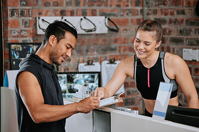 Asian man signing up for gym membership with caucasian trainer. Young coach behind reception pointing and showing where to sign. Two active and fit people standing together in health and fitness club