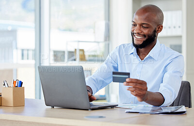 Young happy african american businessman using a credit card and laptop in an office at work alone. One cheerful male business professional making an online purchase with a debit card while sitting at a desk