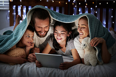Happy caucasian family with two children using digital tablet lying under blanket in the dark at night with their faces illuminated by device screen light. Family of four reading online story or watching movie before bedtime