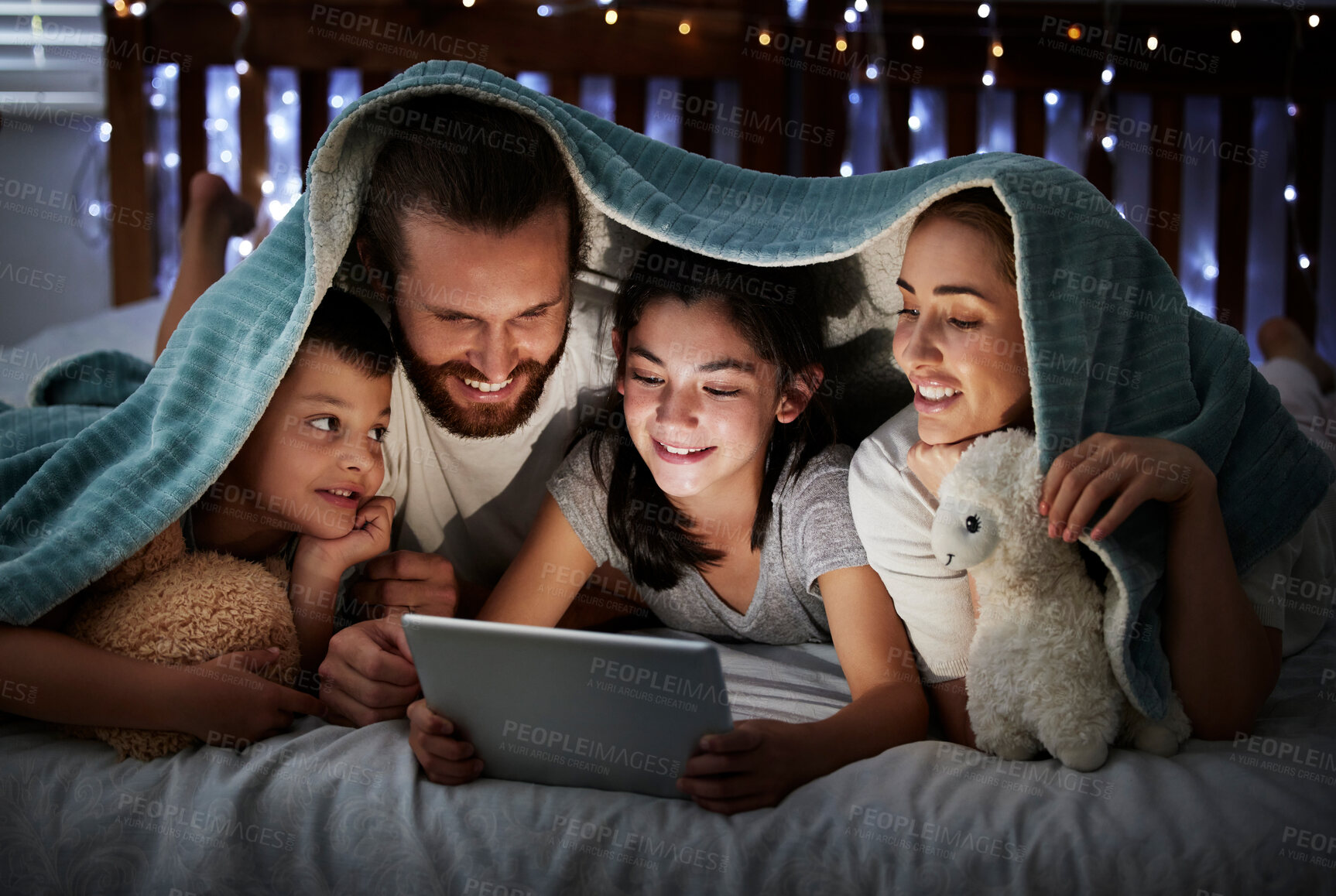 Buy stock photo Happy caucasian family with two children using digital tablet lying under blanket in the dark at night with their faces illuminated by device screen light. Family of four reading online story or watching movie before bedtime