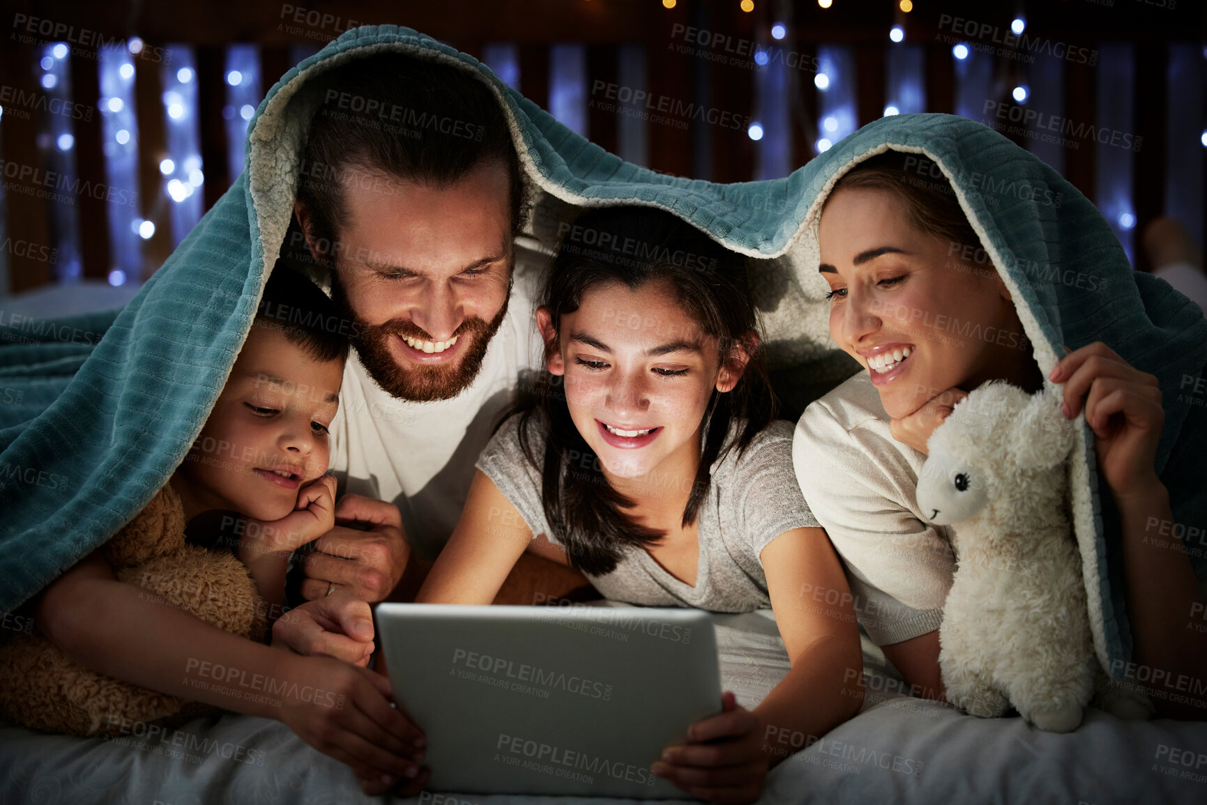 Buy stock photo Happy caucasian family with two children using digital tablet lying under blanket in the dark at night with their faces illuminated by device screen light. Family of four reading online story or watching video before bedtime