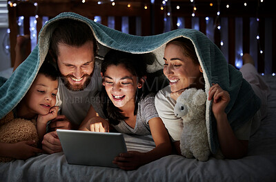 Happy caucasian family with two children using digital tablet lying under blanket in the dark at night with their faces illuminated by device screen light. Family of four reading online story or watching movie before bedtime