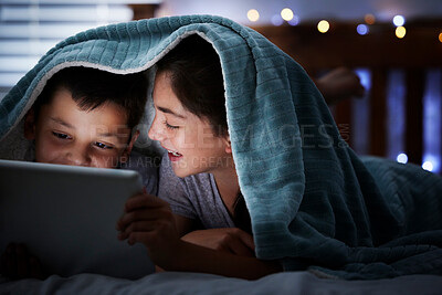 Two caucasian children holding digital tablet while lying under blanket in the dark at night reading online book, watching or playing game before sleeping. Sibling sister and brother lying in bed and faces illuminated by device screen light