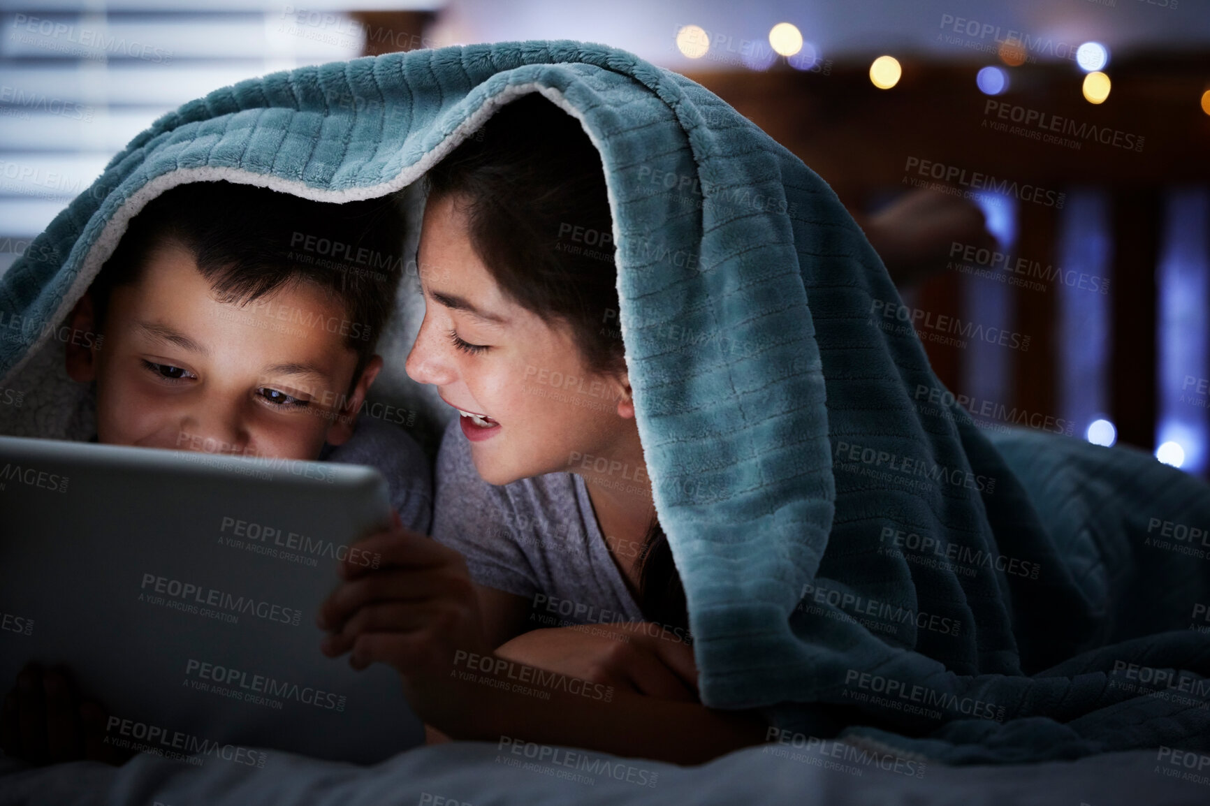 Buy stock photo Two caucasian children holding digital tablet while lying under blanket in the dark at night reading online book, watching or playing game before sleeping. Sibling sister and brother lying in bed and faces illuminated by device screen light