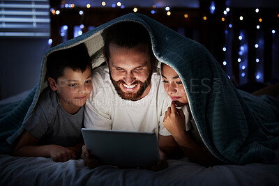 Happy caucasian family single dad with two children using digital tablet lying under blanket in the dark at night with their faces illuminated by device screen light. Father reading online story or watching movie with daughter and son at night