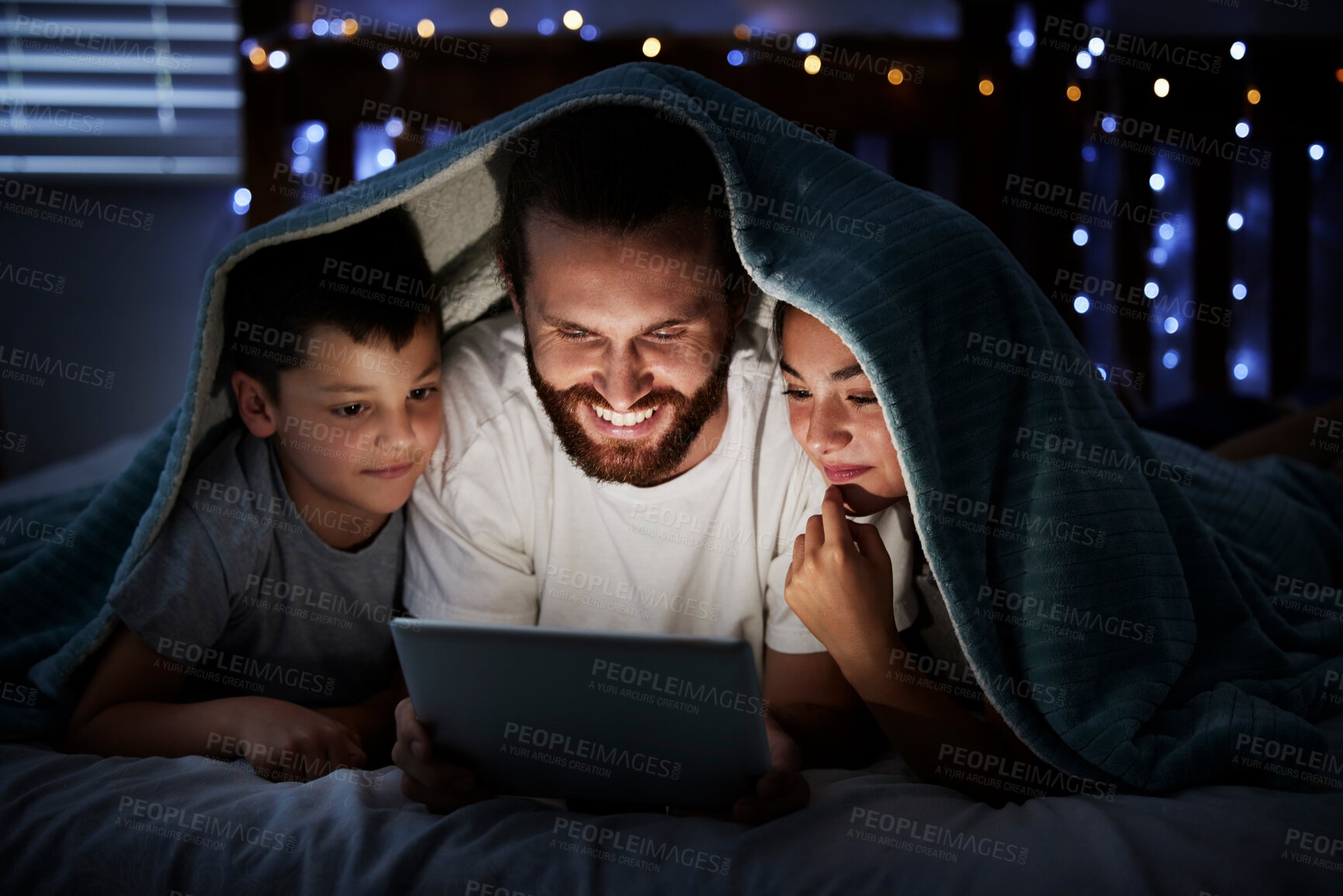 Buy stock photo Happy caucasian family single dad with two children using digital tablet lying under blanket in the dark at night with their faces illuminated by device screen light. Father reading online story or watching movie with daughter and son at night