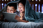 Two kids holding digital tablet while lying under blanket in the dark at night reading online book, watching or playing game before sleeping. Little boy and sister lying in bed and faces illuminated by device screen light