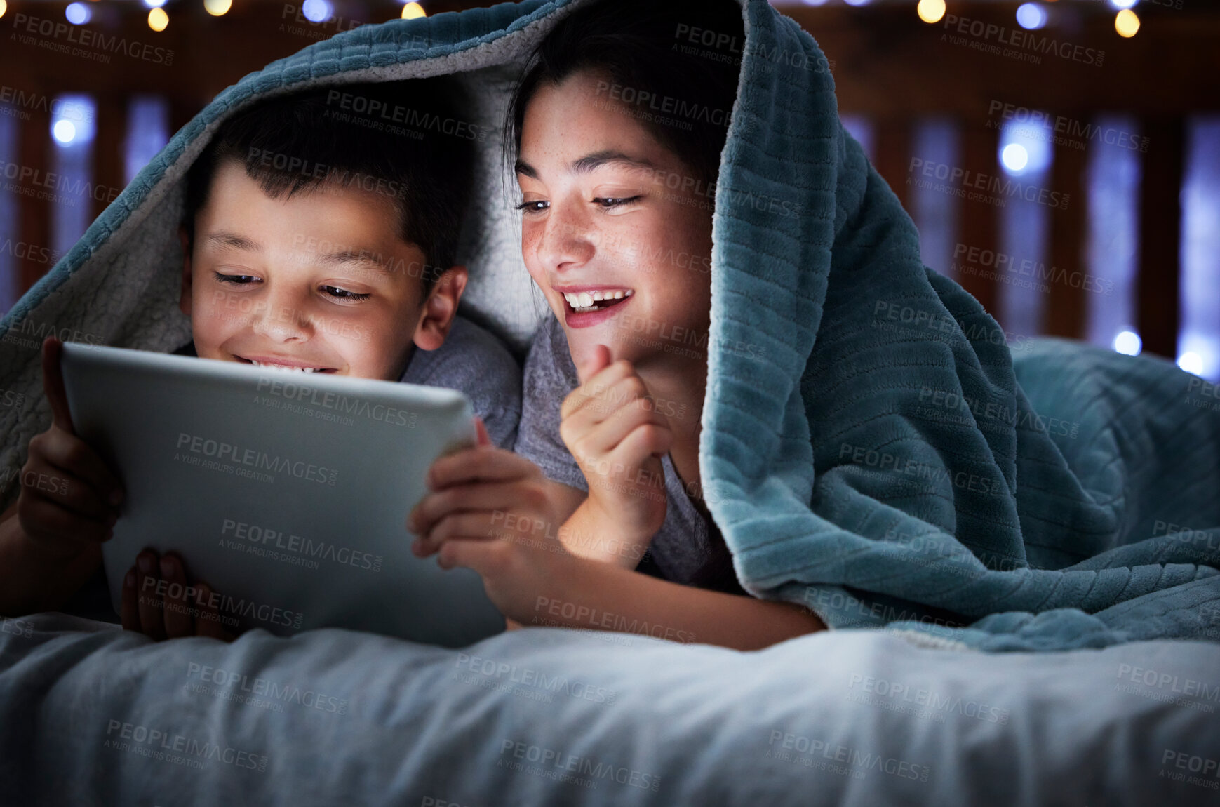 Buy stock photo Two kids holding digital tablet while lying under blanket in the dark at night reading online book, watching or playing game before sleeping. Little boy and sister lying in bed and faces illuminated by device screen light