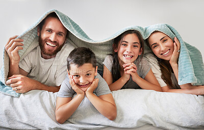 Portrait of happy caucasian family of four lying together on bed with a blanket over their heads. Carefree parents spending free time with their daughter and son over the weekend. Smiling family staying in bed and enjoying a lazy morning