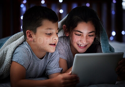Little boy and teenage sibling sister holding digital tablet while lying under blanket in the dark at night reading online book, watching or playing game before sleeping. Two children lying in bed and faces illuminated by device screen light