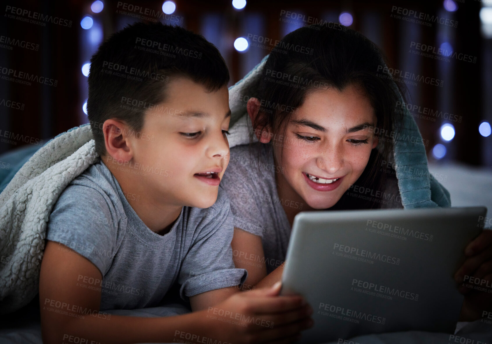 Buy stock photo Little boy and teenage sibling sister holding digital tablet while lying under blanket in the dark at night reading online book, watching or playing game before sleeping. Two children lying in bed and faces illuminated by device screen light