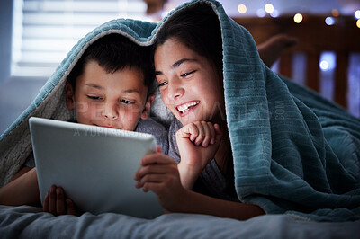 Two kids holding digital tablet while lying under blanket in the dark at night reading online book, watching or playing game before sleeping. Little boy and sister lying in bed and faces illuminated by device screen light