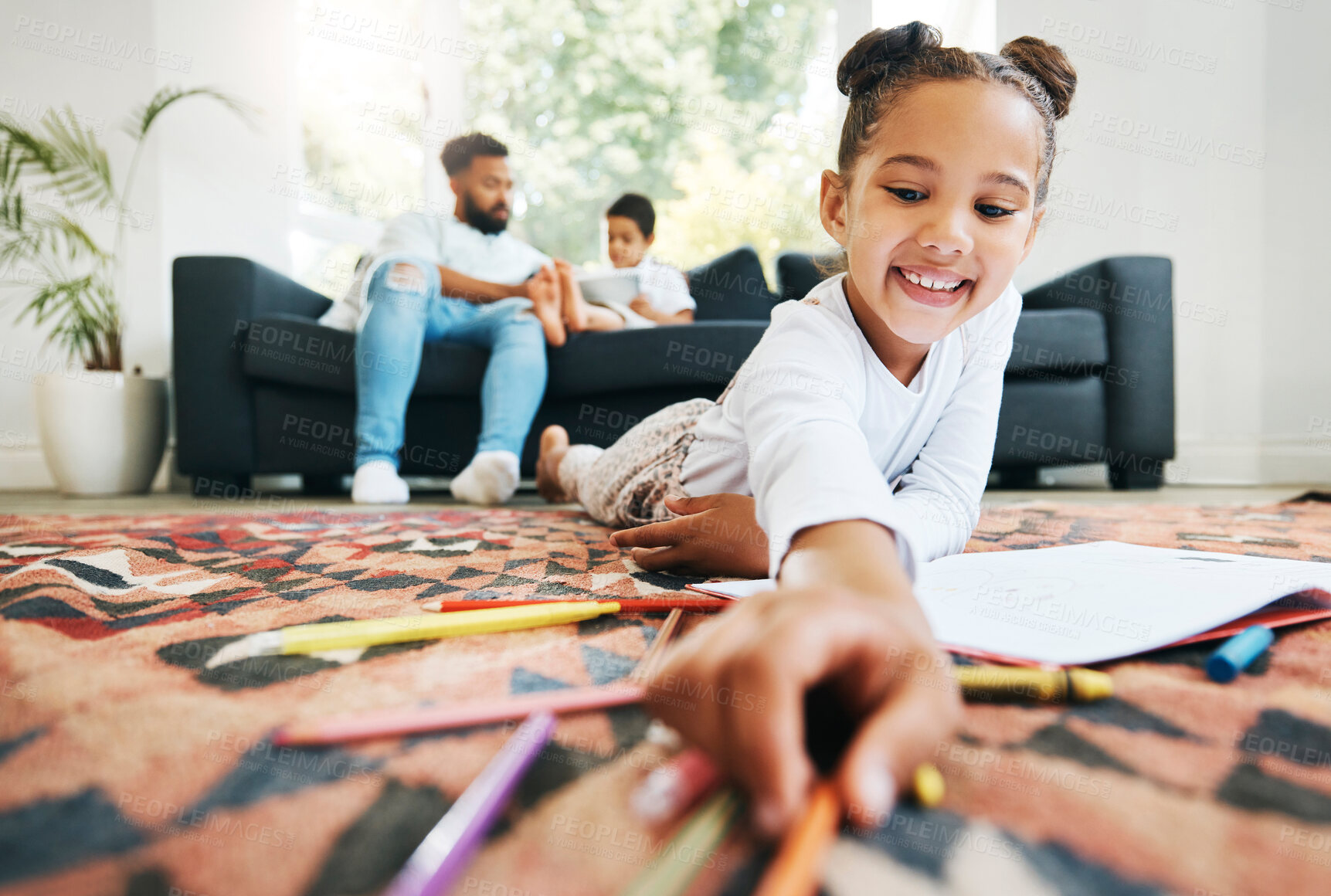 Buy stock photo Little girl drawing with colouring pencils lying on living room floor with her father and brother relaxing on couch. Little girl colouring in during family time at home
