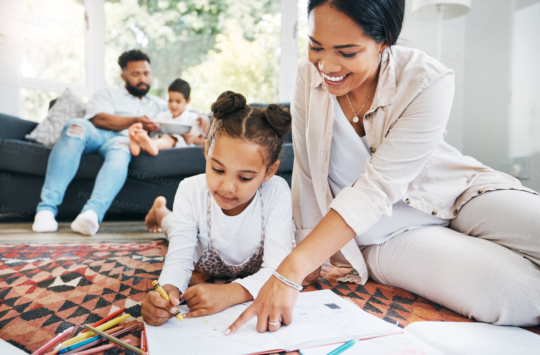 Buy stock photo Little  girl drawing with colouring pencils lying on living room floor with her father and brother relaxing on couch while her mother helps her. Little girl colouring in during family time at home