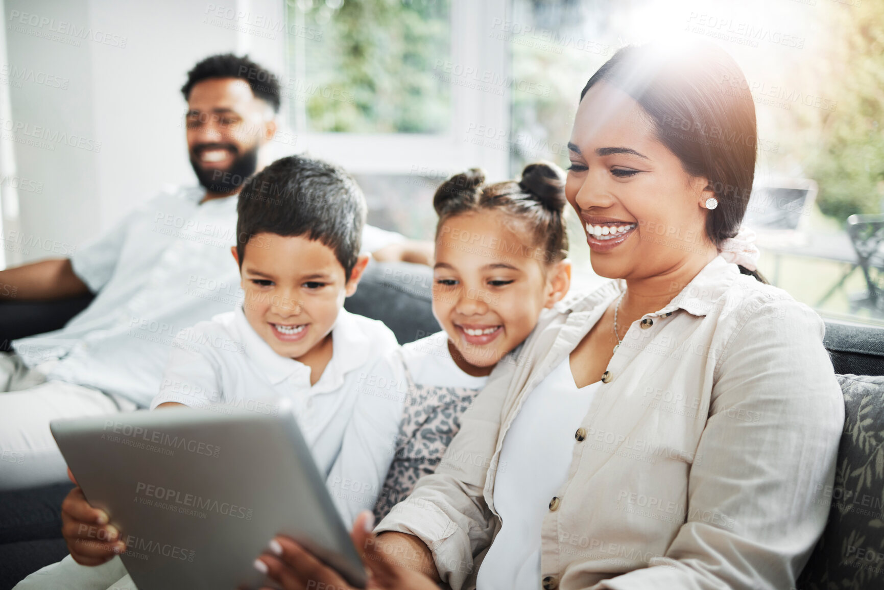Buy stock photo Happy mixed race smiling family of four bonding on a sofa while streaming movies on the internet using a digital tablet at home. Young couple looking cheerful with their daughter and son