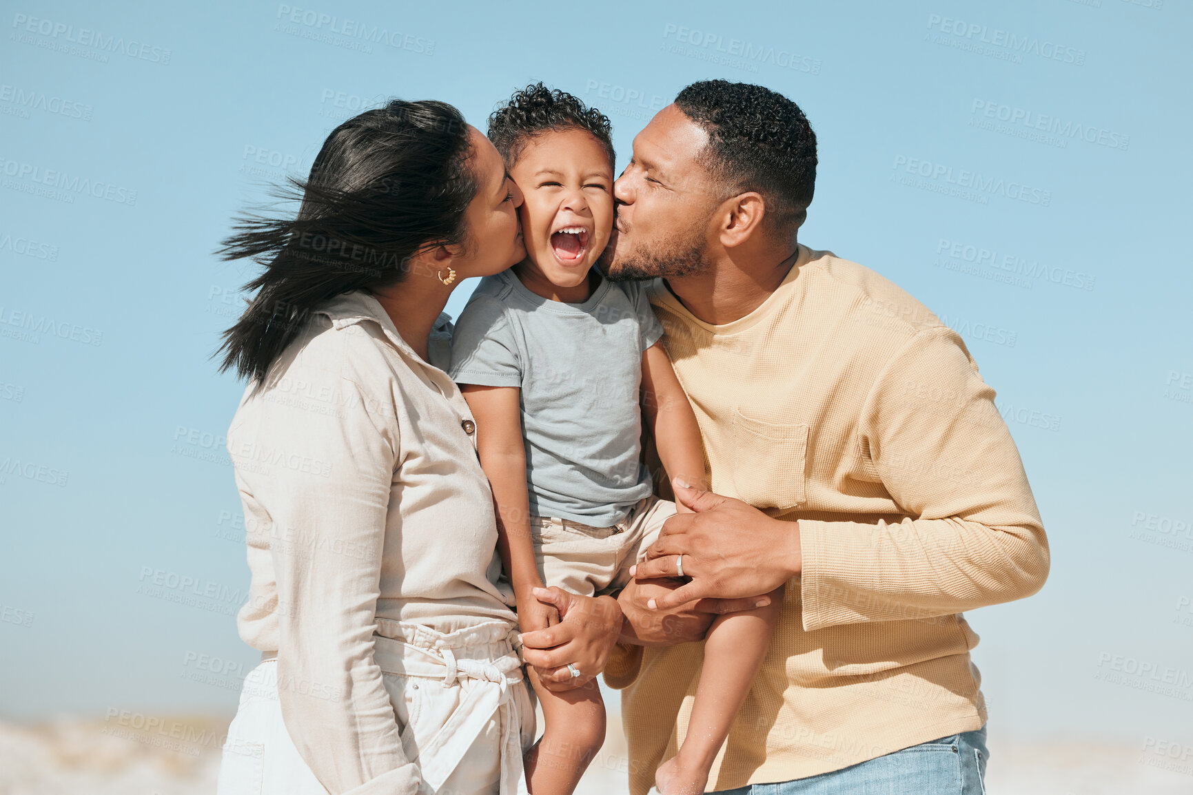 Buy stock photo Happy mixed race family standing on the beach. Loving parents kissing adorable little son on the cheeks showing love and affection while enjoying beach vacation