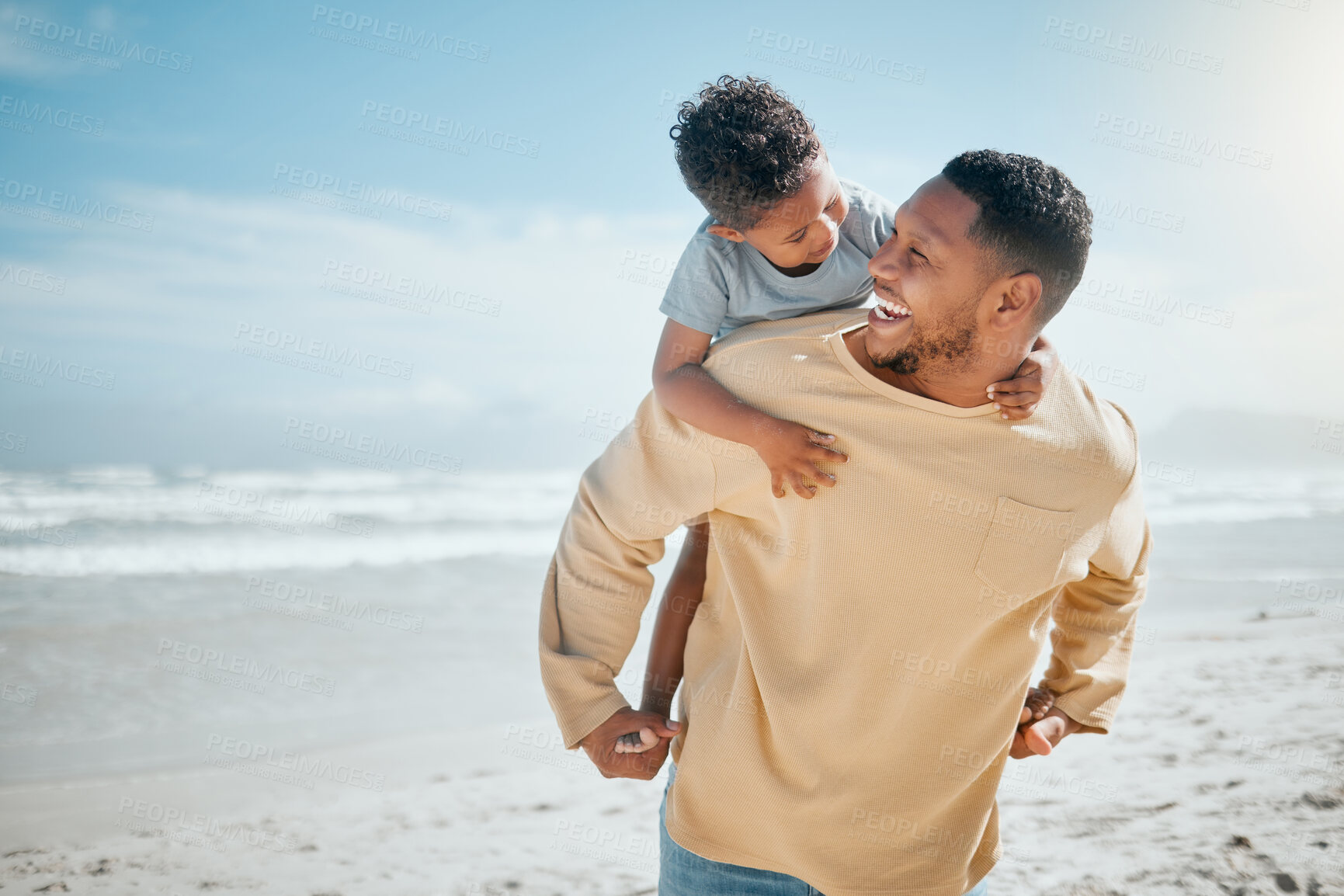 Buy stock photo Cheerful young mixed race father giving his son a piggyback ride on his back having fun and spending time together while on vacation by the beach