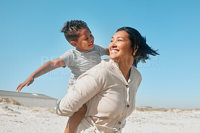 Buy stock photo Cheerful young mixed race mother giving her son a piggyback ride while they look at each other on the beach. Adorable little boy holding his arms outstretched while sitting on his mothers back having fun and enjoying time together while on holiday