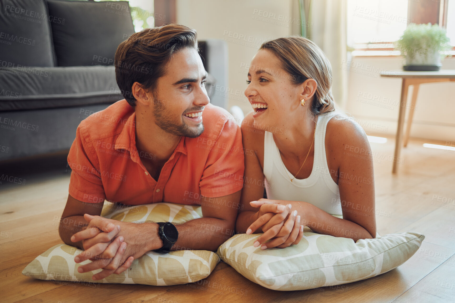 Buy stock photo Loving young caucasian couple looking at each other and laughing while lying on the living room floor with cushions. Couple spending time and happy to be together