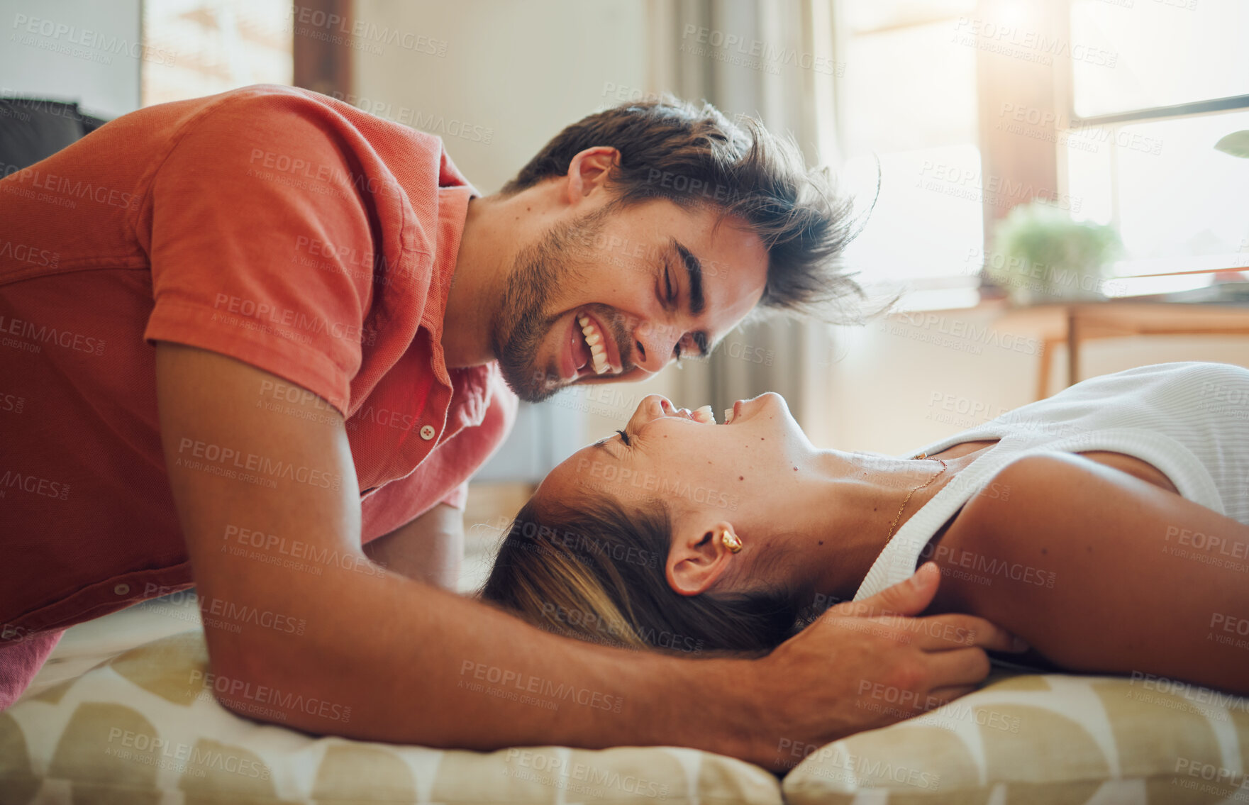 Buy stock photo Happy young woman lying down while her boyfriend leans over her as they laugh together and look into each other eyes. Affectionate couple enjoying romantic intimate moment at home