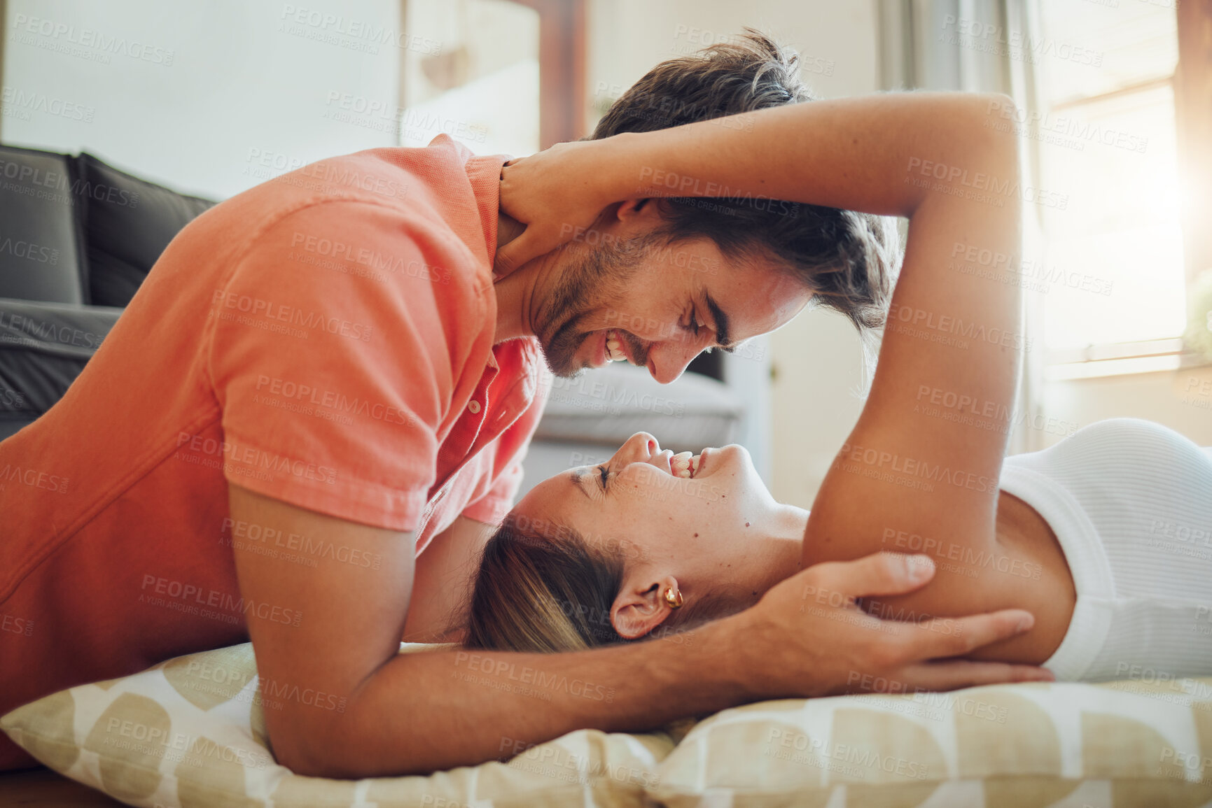 Buy stock photo Happy young woman lying down while her boyfriend leans over her as they laugh together and look into each other eyes. Affectionate couple enjoying romantic intimate moment at home