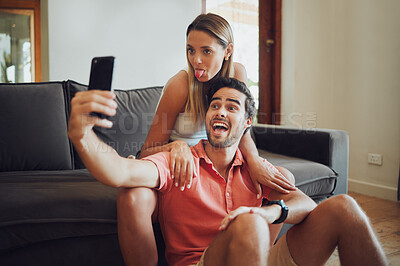Funny romantic couple pulling faces and fooling around while holding a mobile phone and taking a selfie together. Playful man and woman sitting together and taking picture or doing video call together