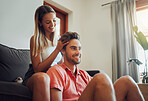 Loving young caucasian couple sitting together at home spending time and happy to be together. Happy young woman sitting on couch while her boyfriend sits between her legs as she plays with his hair. Girlfriend giving head massage
