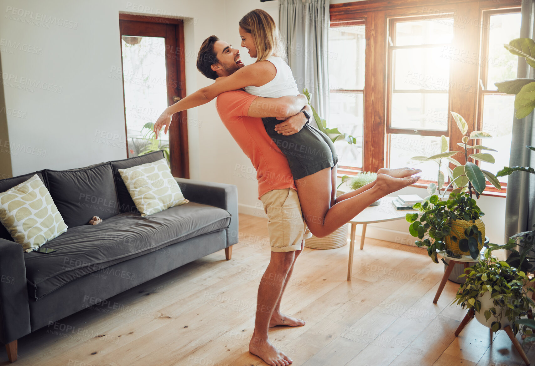 Buy stock photo Happy young boyfriend holding girlfriend in arms as he lifts her up while they look into each others eyes and share intimate moment. Romantic young couple hugging and enjoying passionate dance at home
