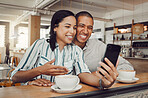 Happy young mixed race couple sitting at table having coffee while looking at something on smartphone in cafe. Loving couple smiling while taking selfie or doing video call on mobile phone while on a date