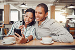 Happy young mixed race couple sitting at table having coffee while looking at something on smartphone in cafe. Loving couple smiling while taking selfie or doing video call on mobile phone while on a date