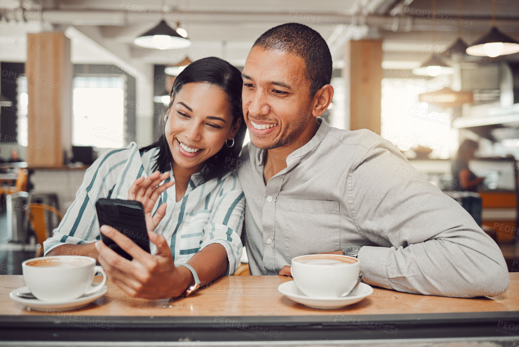 Buy stock photo Happy young mixed race couple sitting at table having coffee while looking at something on smartphone in cafe. Loving couple smiling while taking selfie or doing video call on mobile phone while on a date