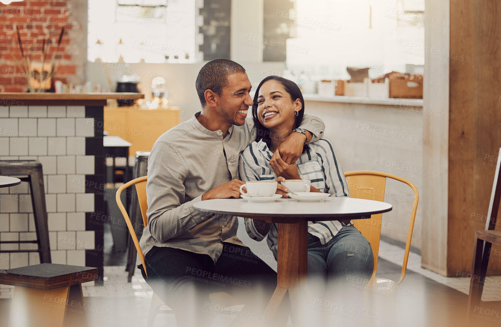 Buy stock photo Loving young man sitting with his arm around his girlfriend while sitting at a table and having coffee in a cafe. Loving mixed race couple talking and laughing while on a date