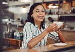 Happy young smiling mixed race woman in casual outfit drinking hot beverage while sitting alone at a table in cozy cafe. Beautiful female looking happy and thoughtful drinking coffee and enjoying her leisure time alone