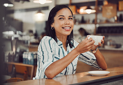 Happy young smiling mixed race woman in casual outfit drinking hot beverage while sitting alone at a table in cozy cafe. Beautiful female looking happy and thoughtful drinking coffee and enjoying her leisure time alone