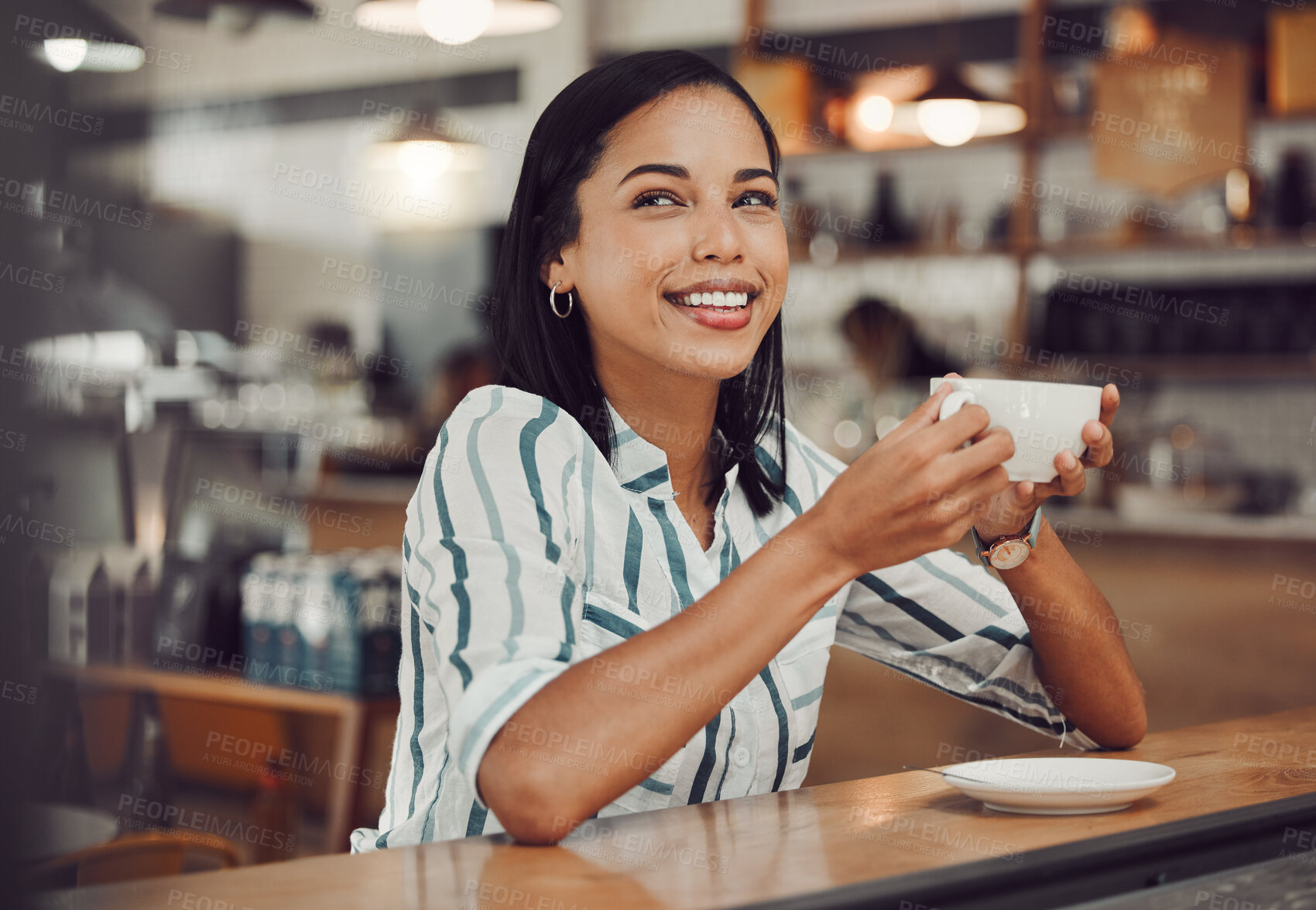 Buy stock photo Happy young smiling mixed race woman in casual outfit drinking hot beverage while sitting alone at a table in cozy cafe. Beautiful female looking happy and thoughtful drinking coffee and enjoying her leisure time alone