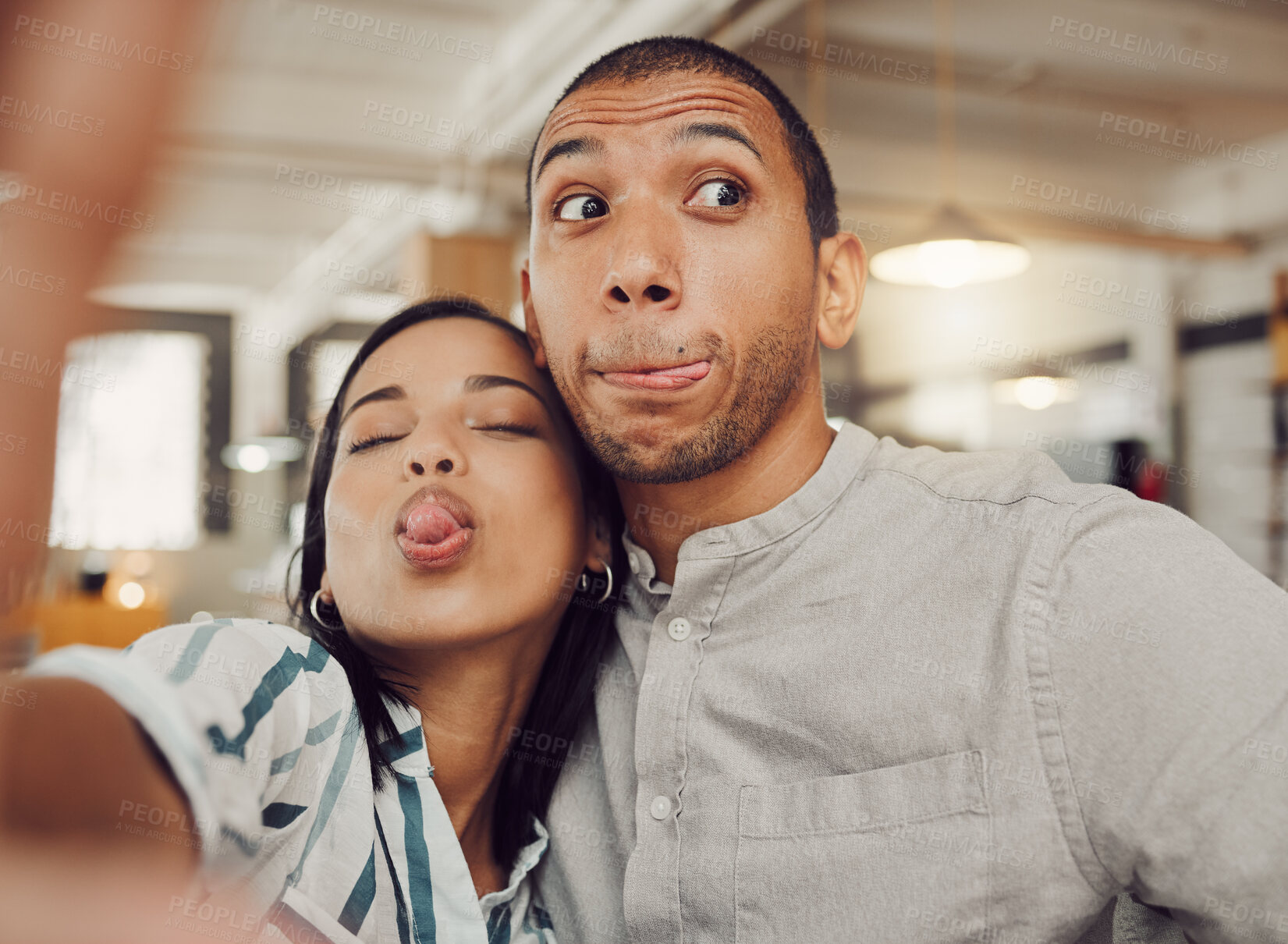 Buy stock photo Close up of funny young woman holding mobile phone while taking a selfie with her boyfriend and making faces in a cafe. Loving mixed race couple being playful while sitting together on a date