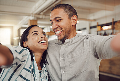 Loving young couple looking at each other and smiling while holding mobile phone to take a selfie in a cafe. Happy mixed race man and woman looking happy while sitting together on a date