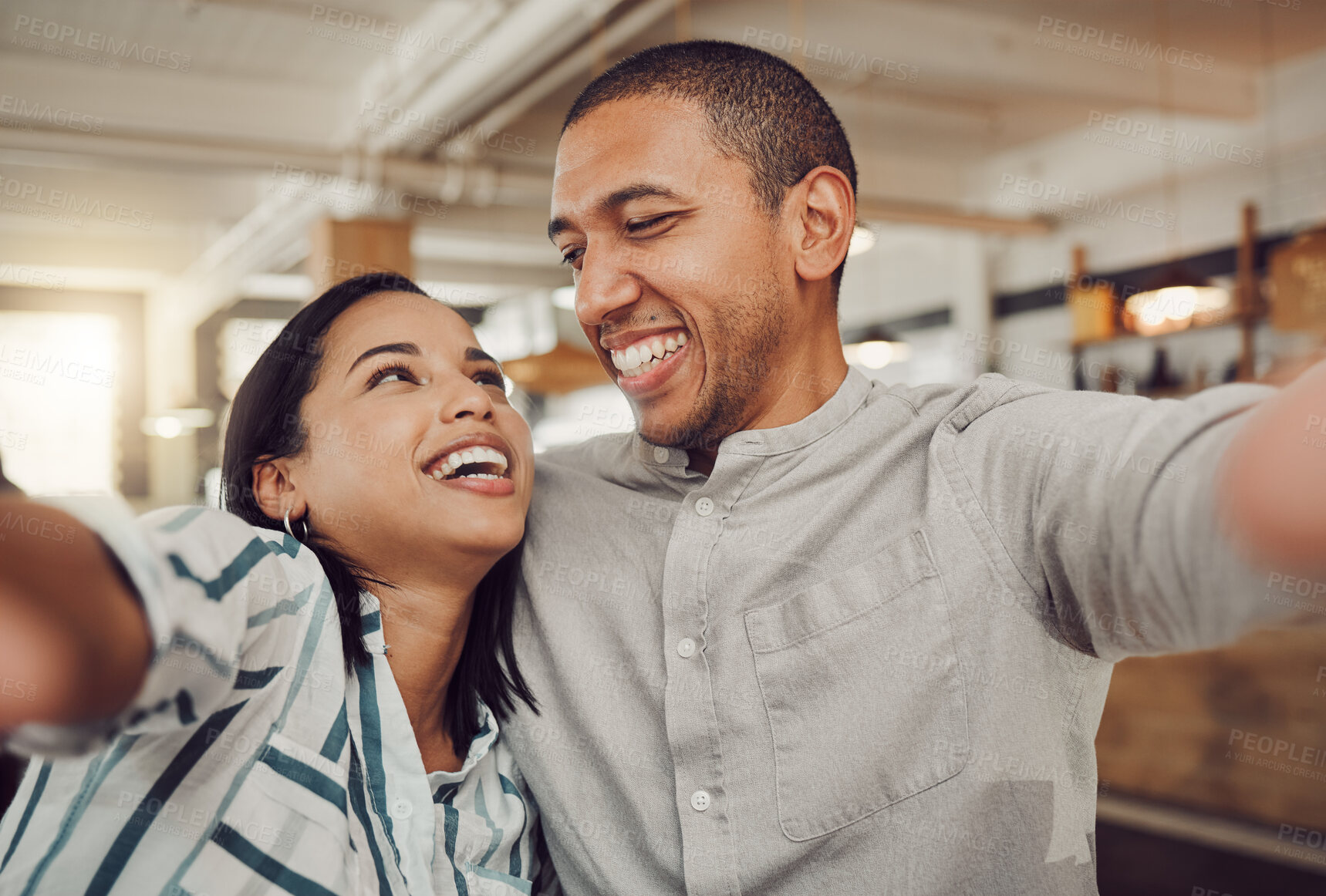 Buy stock photo Loving young couple looking at each other and smiling while holding mobile phone to take a selfie in a cafe. Happy mixed race man and woman looking happy while sitting together on a date