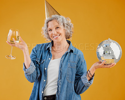 One happy mature caucasian woman holding a disco ball and drinking a glass of white wine while wearing a birthday hat against a yellow background in the studio. Smiling white lady celebrating