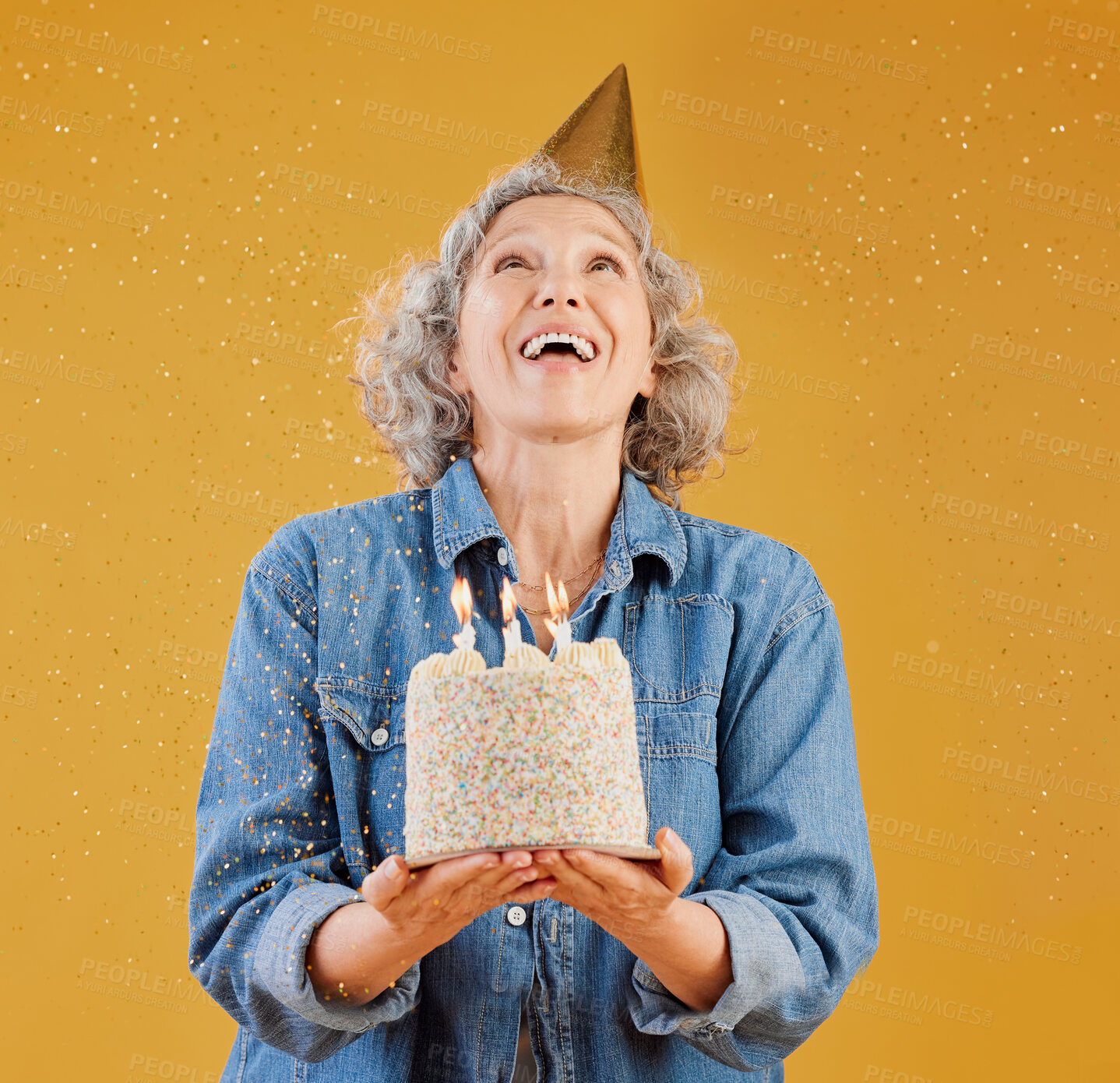 Buy stock photo One happy mature caucasian woman wearing a birthday hat and holding a cake while confetti falls from above against a yellow background in the studio. Smiling white lady celebrating another year