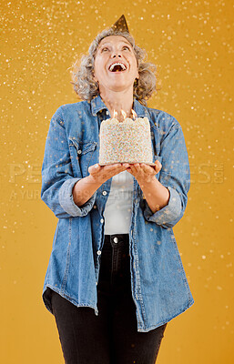 One happy mature caucasian woman wearing a birthday hat and holding a cake while confetti falls from above against a yellow background in the studio. Smiling white lady celebrating another year