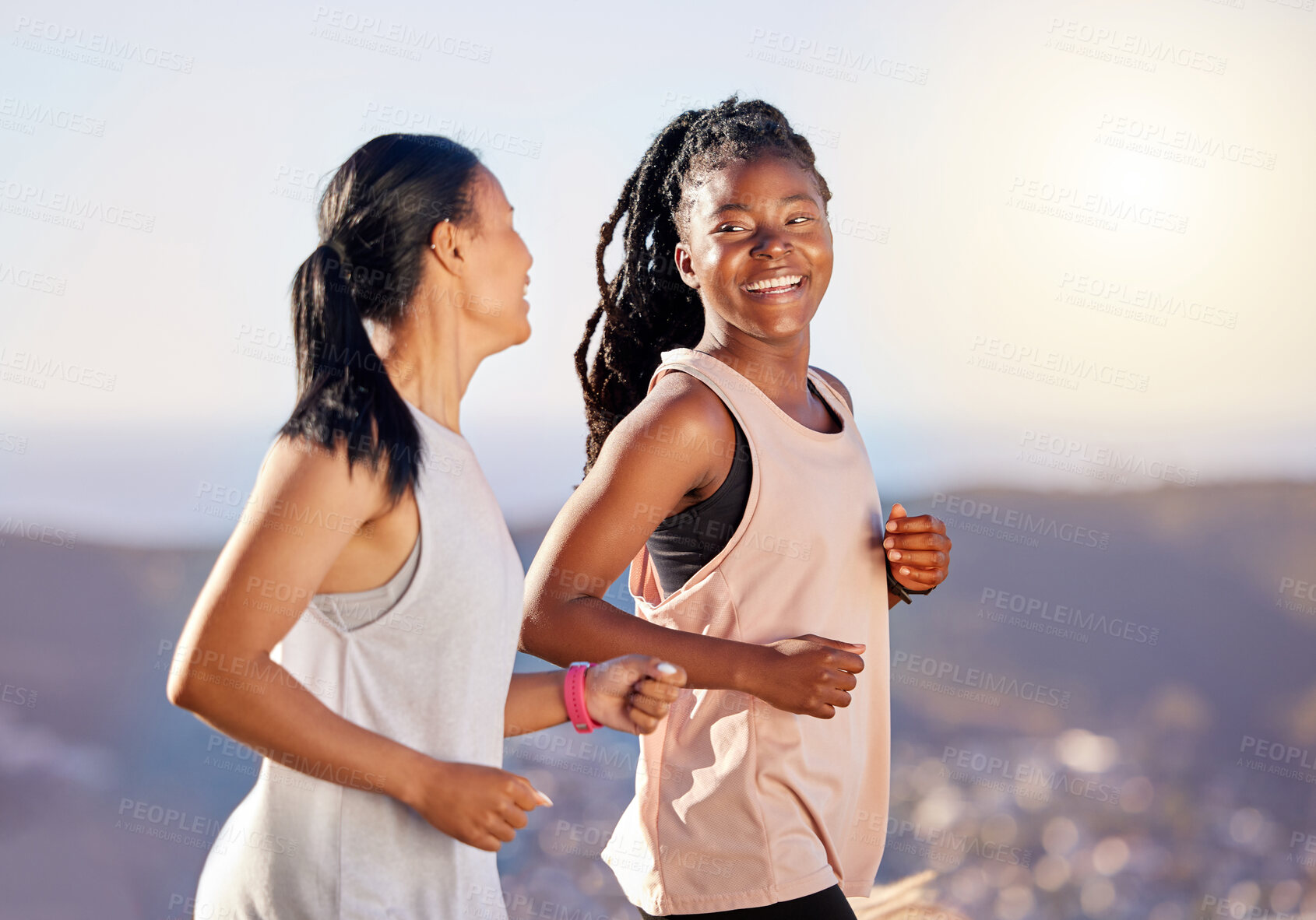 Buy stock photo Two happy young female athletes smiling and talking while out for a run on a mountain road on a sunny day. Energetic young women running outdoors to help their bodies in shape and fit. Two diverse female friends exercising together