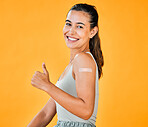 Mixed race covid vaccinated woman with arm plaster, showing thumbs up sign and symbol. Portrait of hispanic model isolated against yellow studio background with copyspace. Endorsing corona vaccine