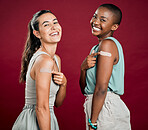 Covid vaccinated African american and mixed race women showing, pointing at arm plaster. Two happy people isolated on red studio background with copyspace. Black woman and hispanic with corona vaccine